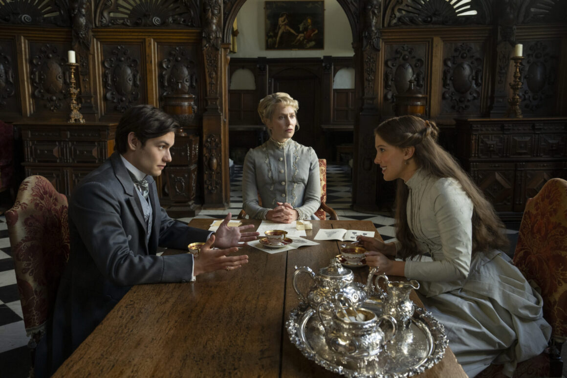 "Three people in Victorian-era clothing sit around a wooden table with silver tea set in an ornate, wood-paneled room, engaged in conversation."
